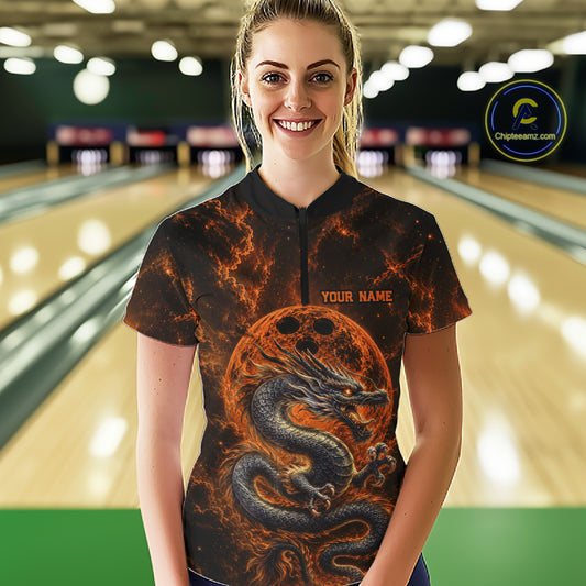 Front view of a happy female model in a bowling alley wearing an orange custom quarter-zip bowling shirt featuring an ancient dragon, flames, and a moon-inspired bowling ball design.
