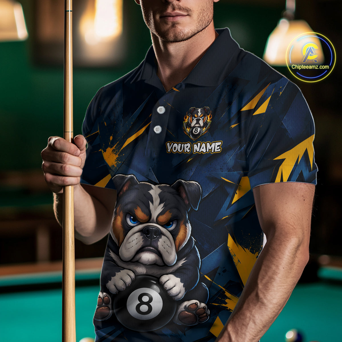 A front view of a custom blue billiards polo shirt worn by a male model holding a pool cue. The shirt features a funny cartoon bulldog with its paw on an 8-ball on the front. A 'YOUR NAME' text placeholder is positioned below a crossed pool cues icon. The background is a blurred billiards room with a green pool table.