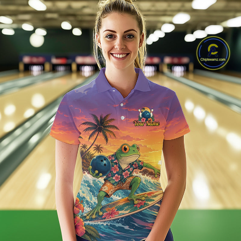 A woman stands in a bowling alley wearing a purple short-sleeve polo shirt with a colorful graphic print of a frog surfing on a wave at sunset, holding a bowling ball. The background shows blurred bowling lanes.