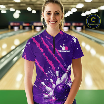 A woman stands in a bowling alley wearing a purple short-sleeve bowling shirt with a black zipper collar and a bowling-themed graphic print featuring white bowling pins and balls. The background shows blurred bowling lanes.
