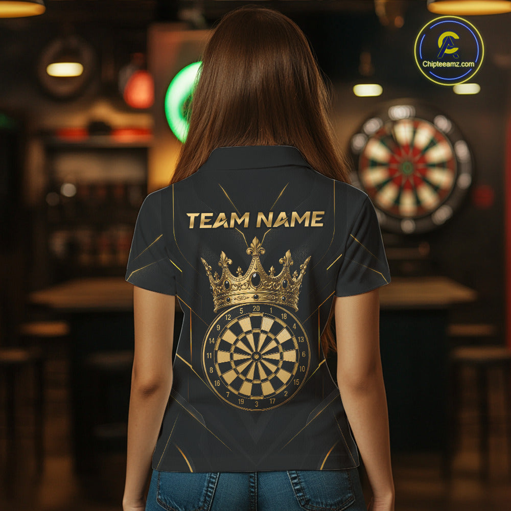 A woman viewed from behind wearing a dark navy or black short-sleeve polo shirt with a large gold crown and dartboard graphic on the back, along with the text 'TEAM NAME'. The background is a blurred bar or dartboard room with another dartboard visible.