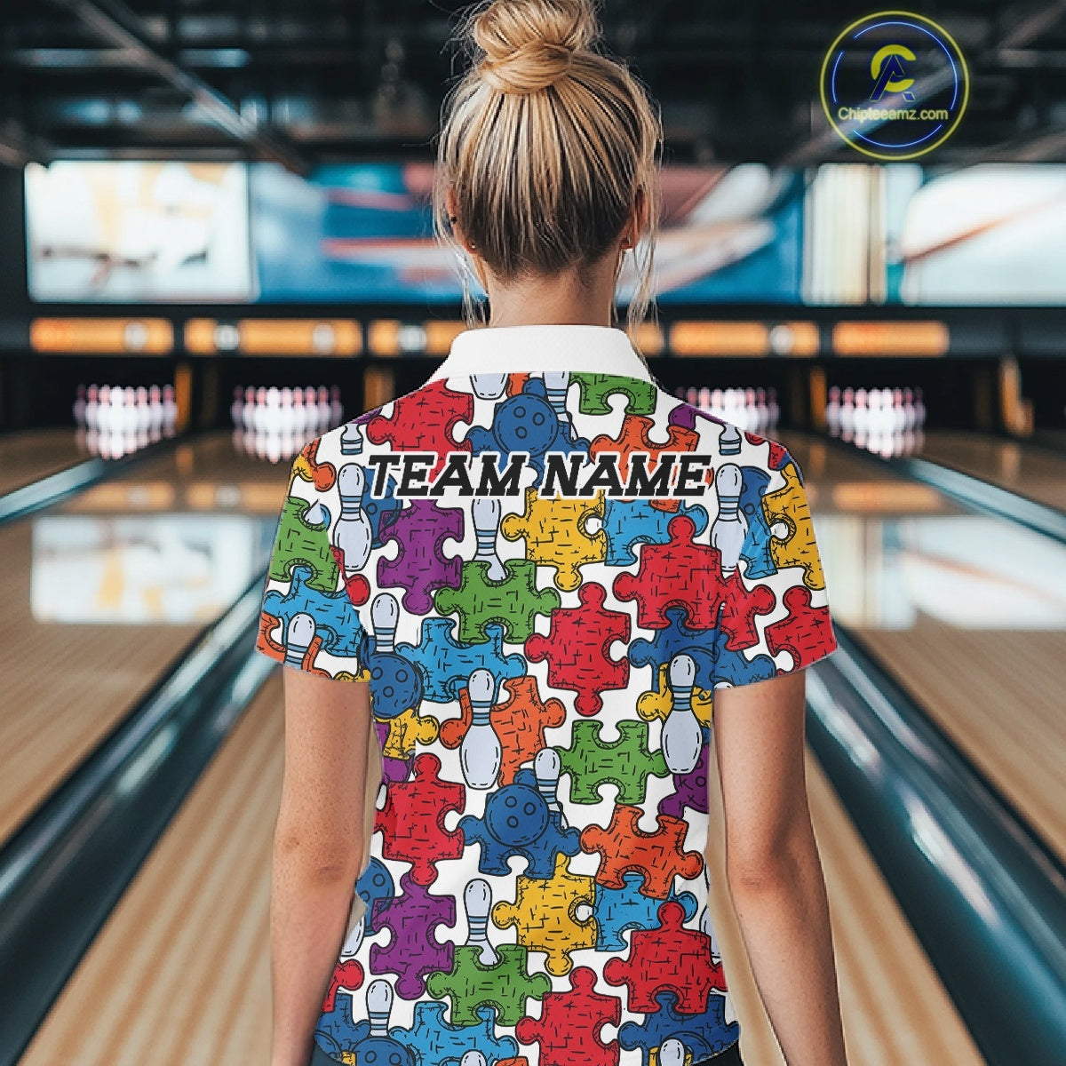 A woman viewed from behind wearing a short-sleeve collared shirt with a colorful puzzle-piece pattern and the text 'TEAM NAME' printed across the upper back. She is standing in a bowling alley with bowling lanes visible in the background.