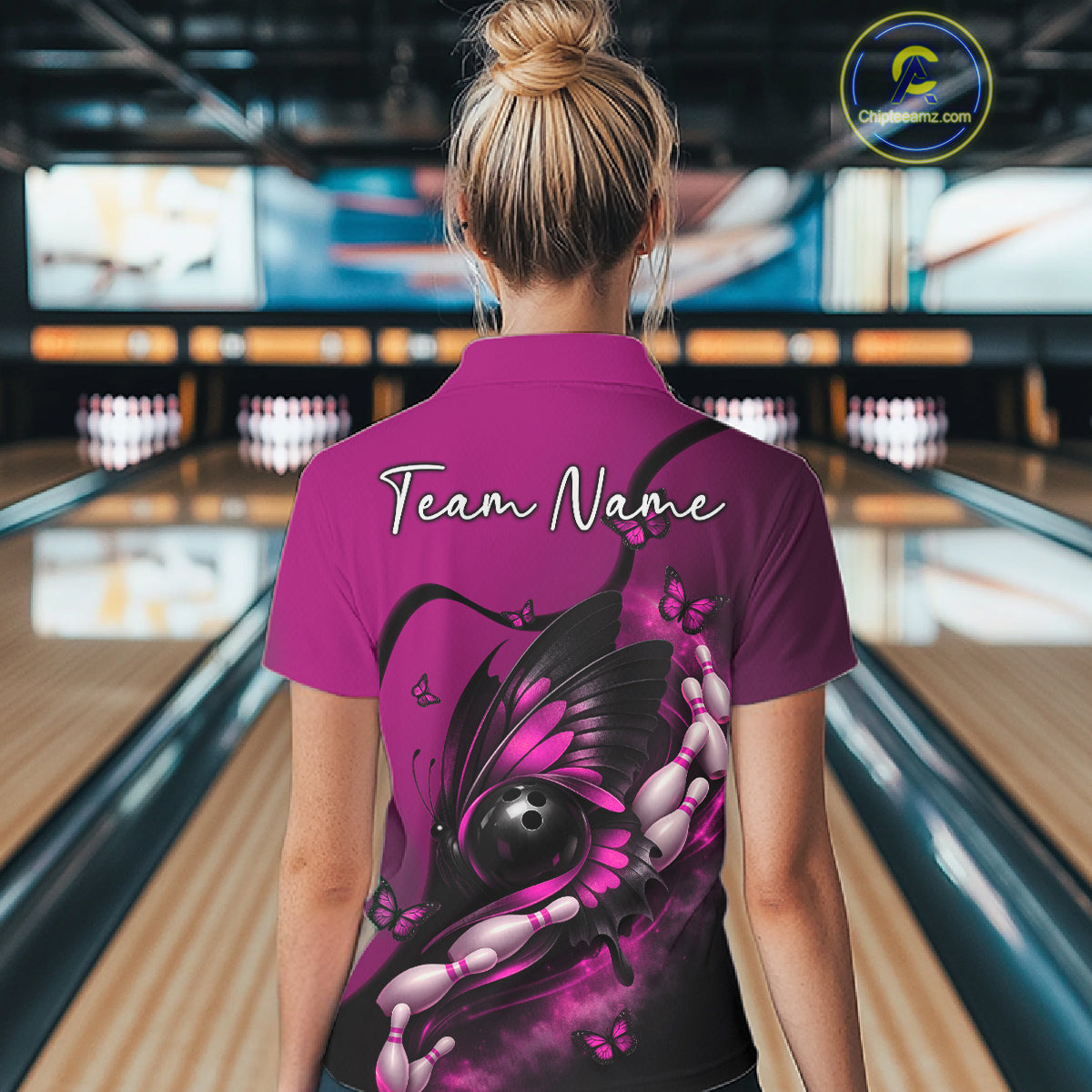 Back view of a female model in a bowling venue wearing a pink and black polo shirt featuring a butterfly and bowling-themed graphics.