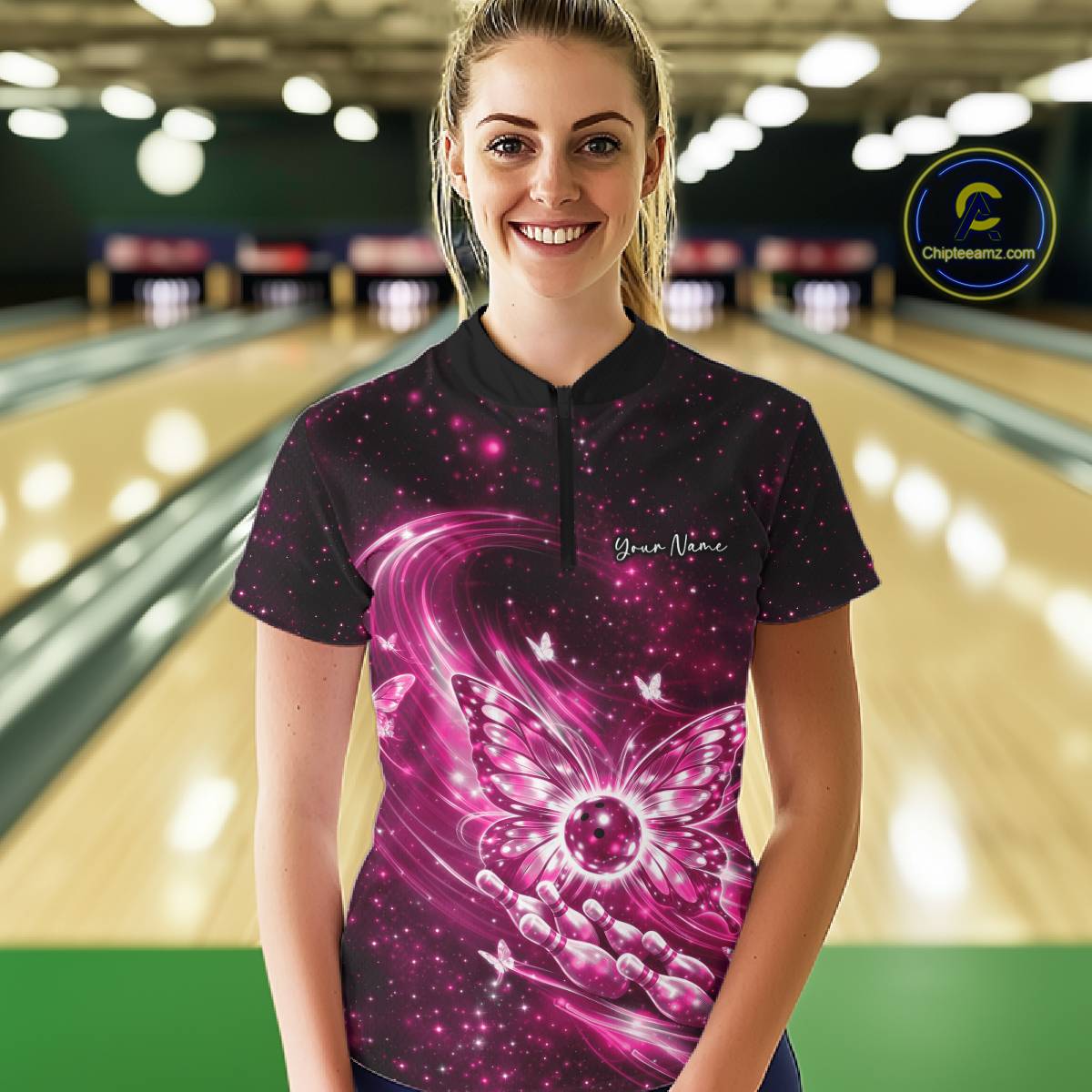 Female model shown from the front in a bowling center wearing a pink quarter-zip shirt with luminous butterfly wings and bowling ball graphics.