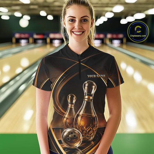 A woman stands in a bowling alley wearing a black short-sleeve quarter zip shirt with a gold and brown bowling-themed graphic print featuring bowling pins that resemble wine bottles and a ball, with 'YOUR NAME' printed on the chest. 