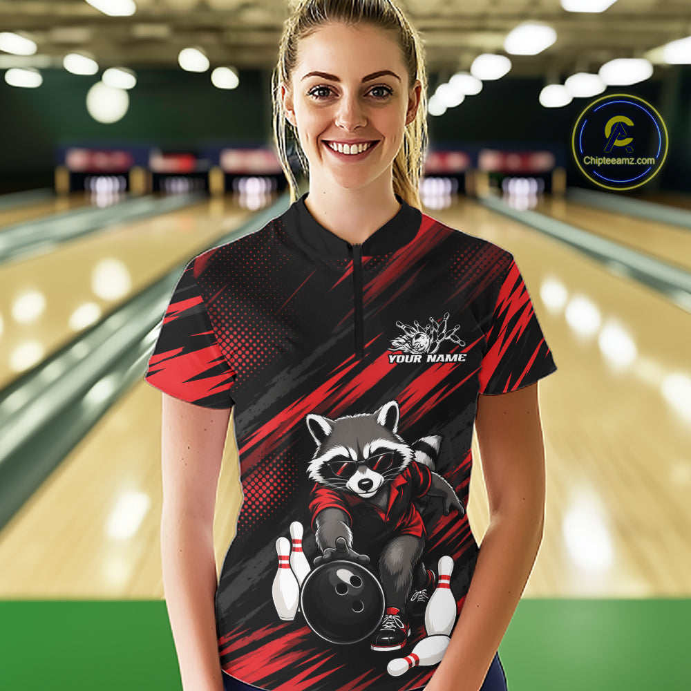 A woman stands in a bowling alley wearing a custom short-sleeve bowling shirt. The shirt features a black and red abstract background with a cartoon raccoon holding a bowling ball and pins, and a white text placeholder 'YOUR NAME'.