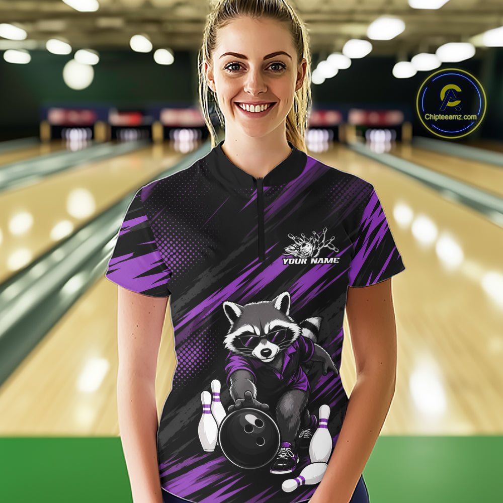A woman stands in a bowling alley wearing a custom short-sleeve quarter zip bowling shirt. The shirt features a black and purple abstract background with a cartoon raccoon holding a bowling ball and pins, and a white 'YOUR NAME' placeholder logo on the chest.
