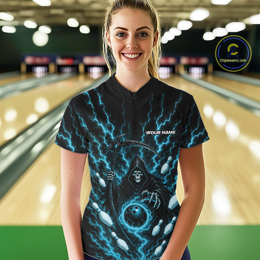 A woman wearing a black short-sleeve quarter zip bowling shirt with a blue lightning and grim reaper design, standing in a bowling alley with blurred lanes in the background.