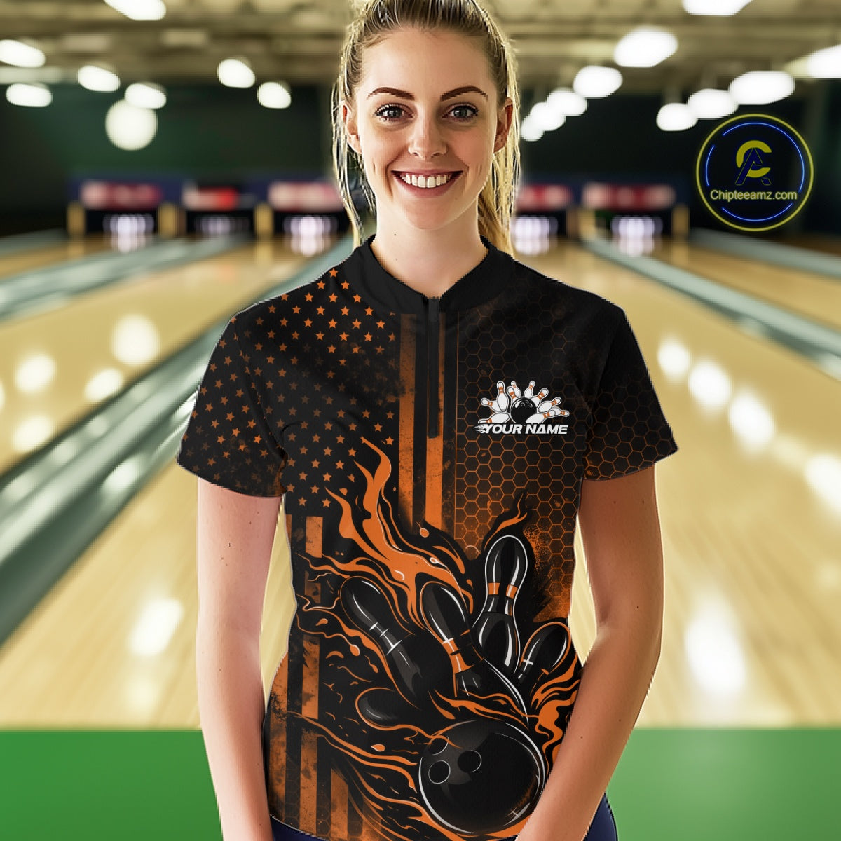 Female model at a bowling lane wearing a black and orange bowling quarter-zip shirt with fiery bowling strike graphics and flag-inspired styling.
