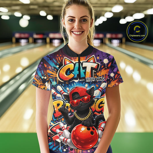 Female model wearing a colorful bowling quarter-zip shirt at a bowling lane with graffiti-style black cat artwork, sunglasses, cap, and bowling ball graphic.