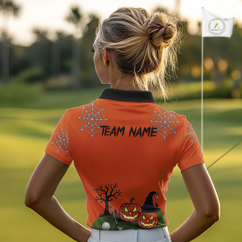 A woman on a golf course seen from behind, wearing a bright orange short-sleeve polo shirt with a black collar. The shirt features a Halloween-themed graphic print on the back with spiderwebs, jack-o'-lanterns, a witch hat, and the text 'TEAM NAME'.