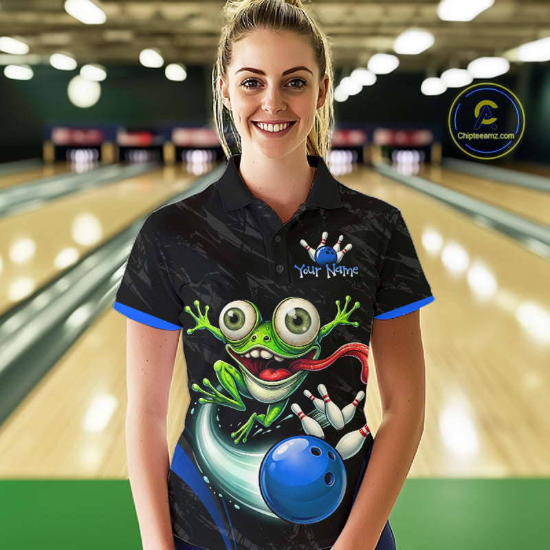 A woman stands in a bowling alley wearing a black short-sleeve polo shirt with a colorful cartoon frog and bowling ball graphic on the front. The shirt has blue trim on the collar and sleeve cuffs, and the words 'Your Name' are printed on the chest as a customizable placeholder.