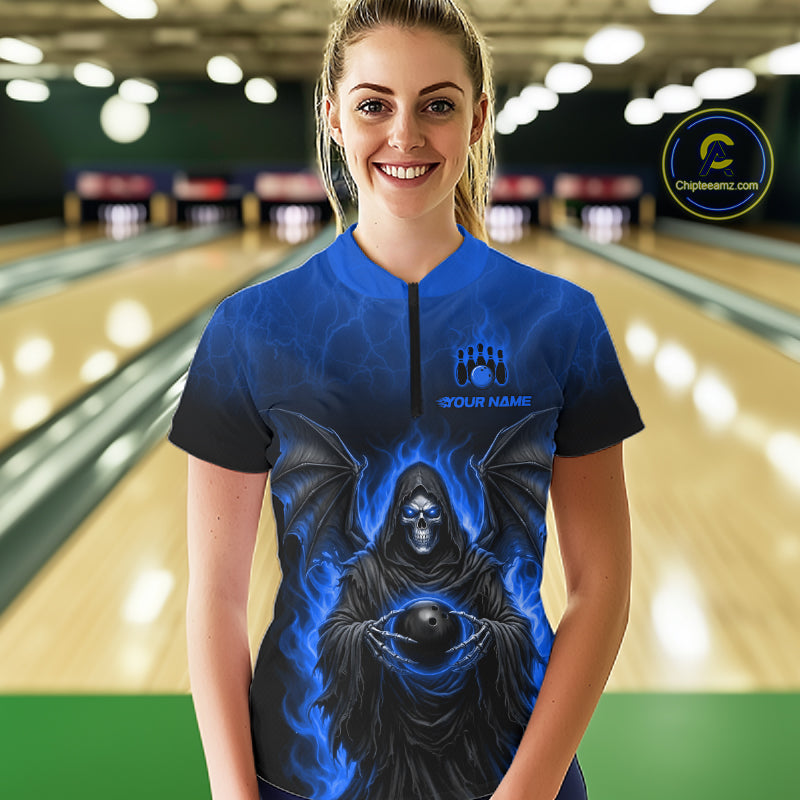 A woman stands in a bowling alley wearing a short-sleeve zip-front bowling shirt with a blue and black grim reaper graphic holding a bowling ball. The shirt has a blue gradient background with a black lower section and a small white 'YOUR NAME' text with a bowling pin icon above it.