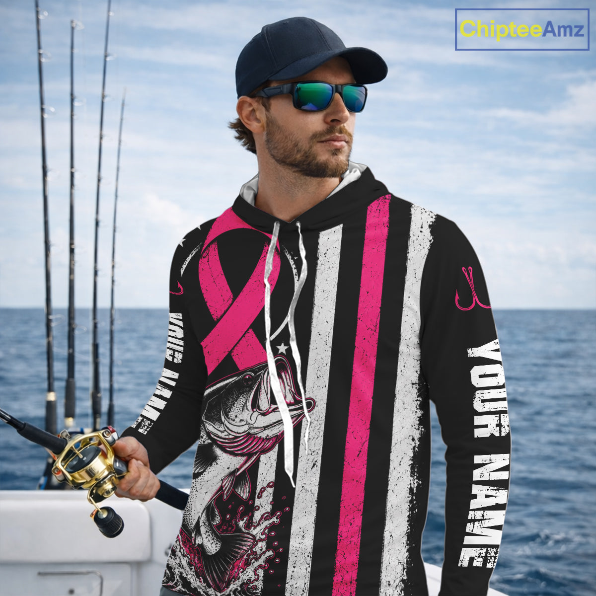 A man on a boat wearing a black custom hoodie with pink and white stripes, a fish graphic, and white text reading 'YOUR NAME'. He is also wearing a dark cap and sunglasses, with fishing rods and blue ocean water in the background.