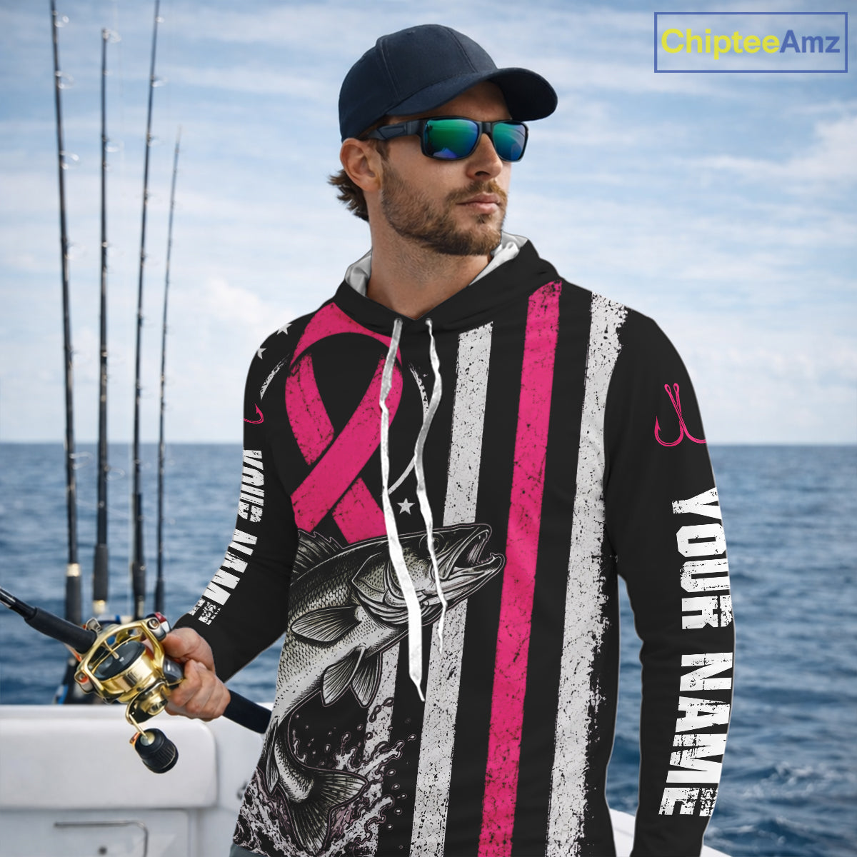 A man on a boat wearing a black custom fishing hoodie with a pink ribbon design, a bass graphic, and vertical white text reading 'YOUR NAME'. He is also wearing a dark cap and sunglasses, holding a fishing rod.