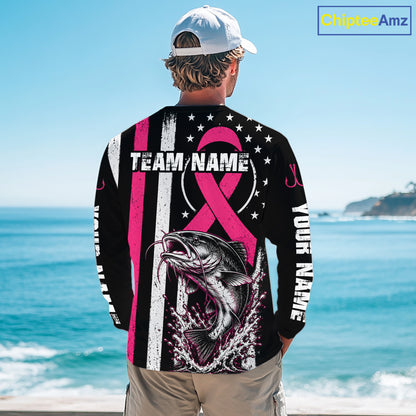 A man stands on a beach with his back to the camera wearing a black long-sleeve shirt with a pink and white patriotic fishing design, including a fish graphic, stars, stripes, and customizable text placeholders.
